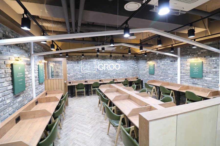 회색 벽돌 벽에 GROO 로고가 있고 나무색 책상과 초록 의자가 놓인 스터디카페 열람실