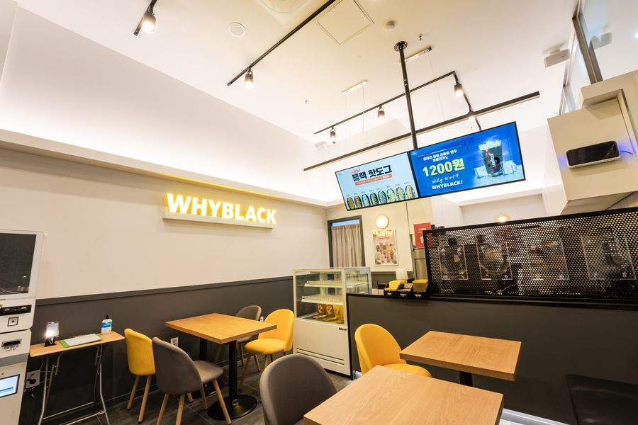 노란 의자와 나무색 테이블, 벽의 WHYBLACK 사인이 보이는 작은 카페 내부